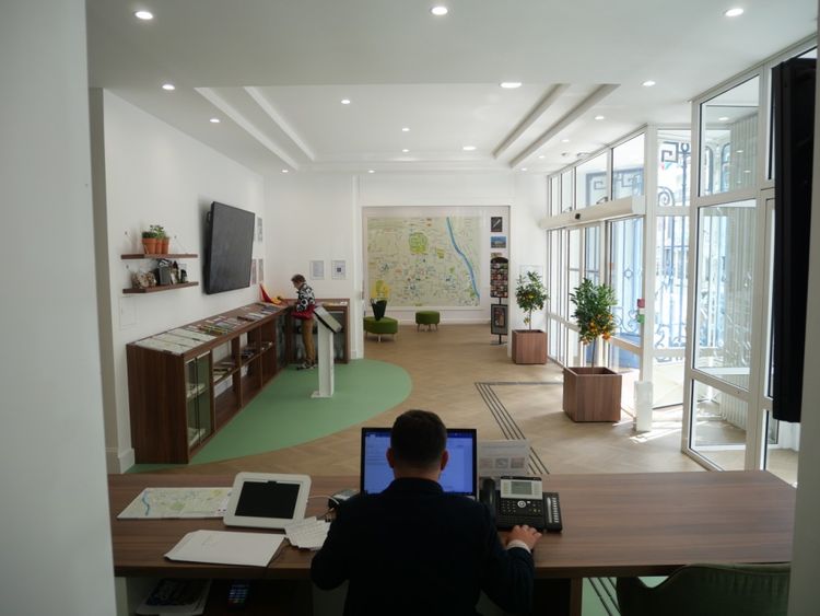 L'entrée de l'Office de Tourisme vu depuis la banque d'accueil avec un homme qui travaille sur ordinateur, une personne qui regarde des brochures ,et l'espace détente au fond avec plan mural